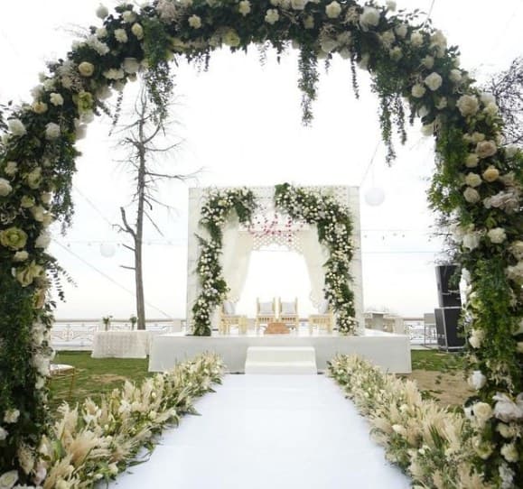 View through a round floral arch down a white aisle to a white mandap