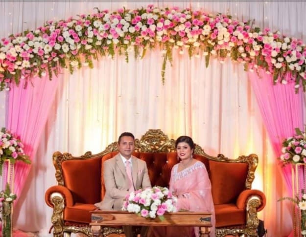 Couple on a carved red sofa under an arch of pink and white flowers with pink side drapes