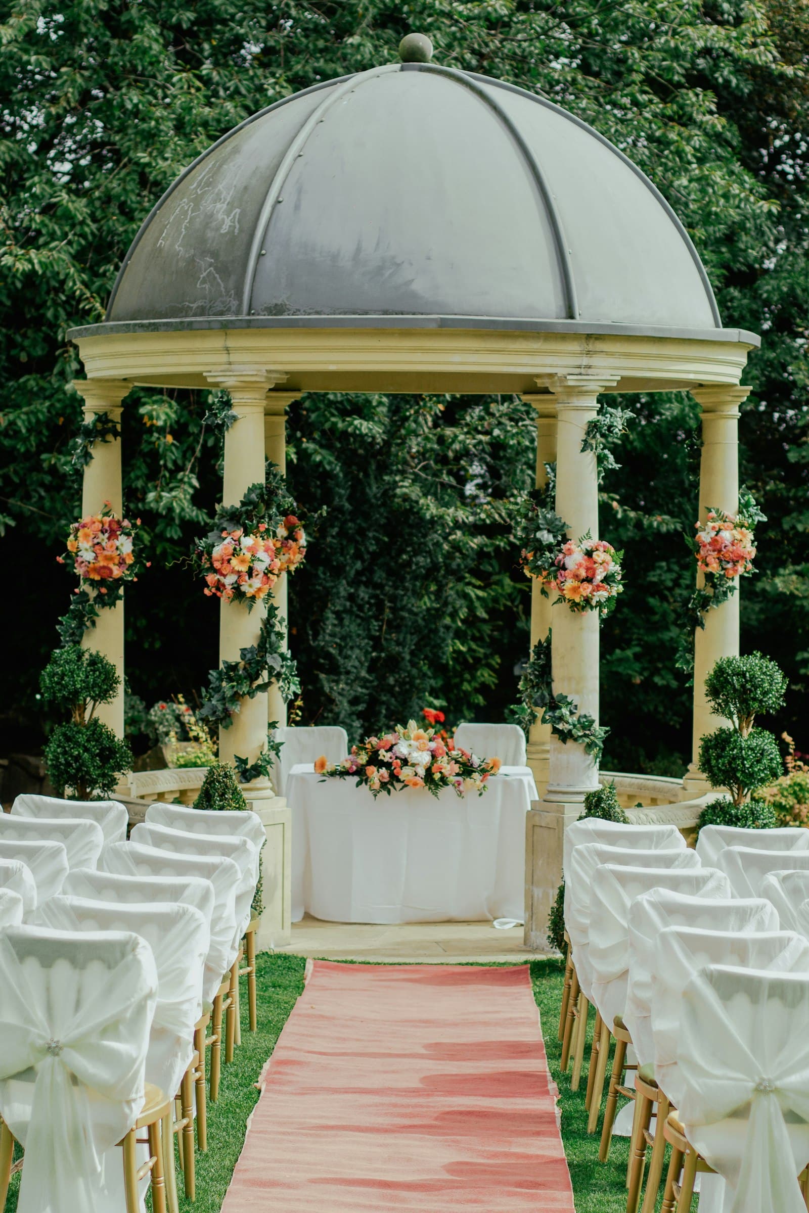 Garden gazebo wrapped in coral florals with white-draped chairs along a pink aisle