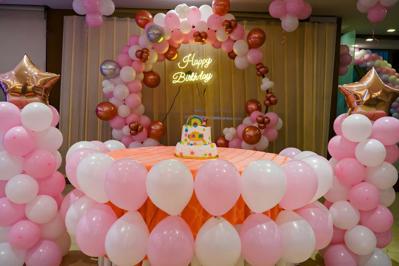 Birthday setup with a pink and rose-gold balloon arch, a neon Happy Birthday sign and a tiered cake