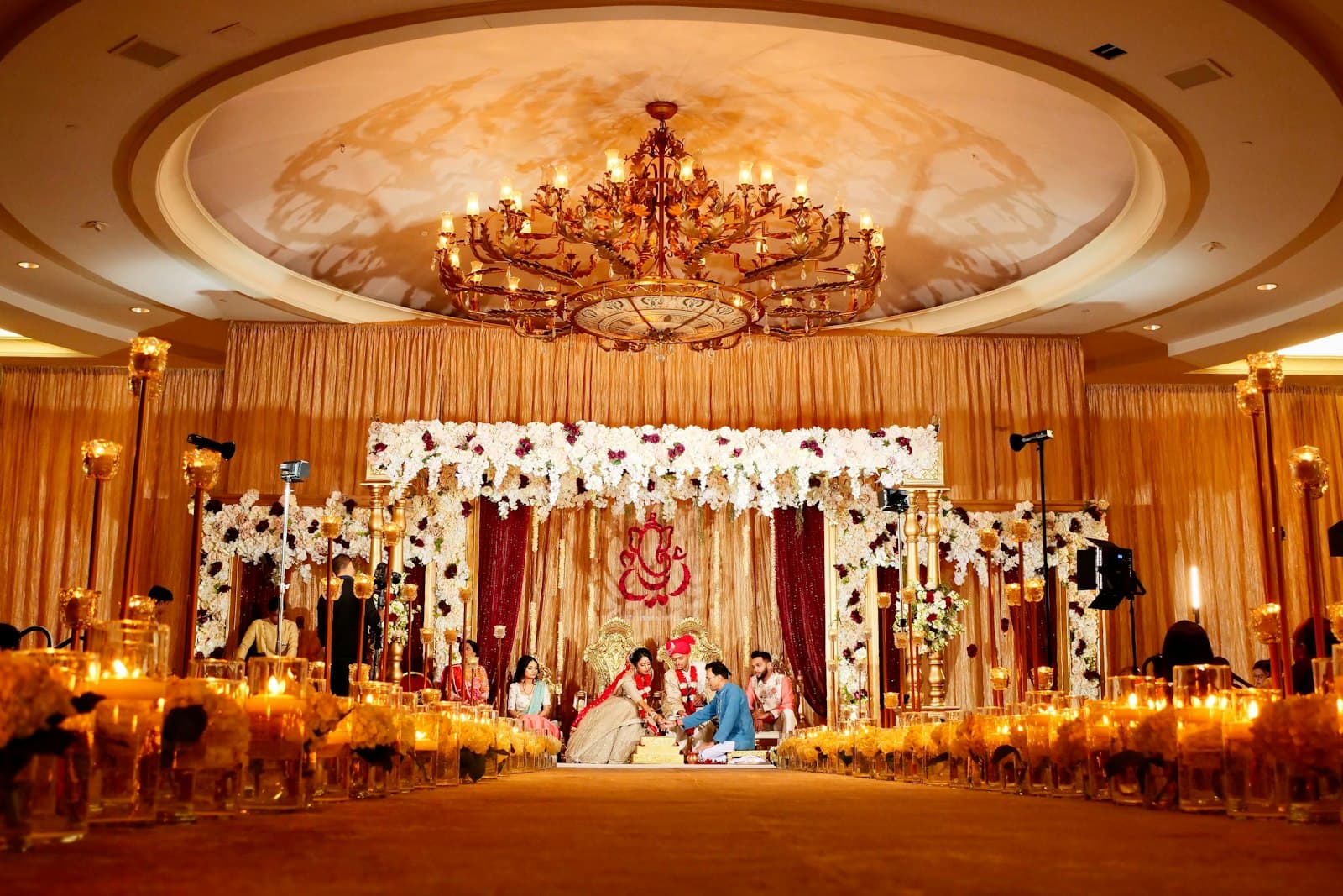 Couple seated for a wedding ritual under a white floral mandap in a candle-lit ballroom